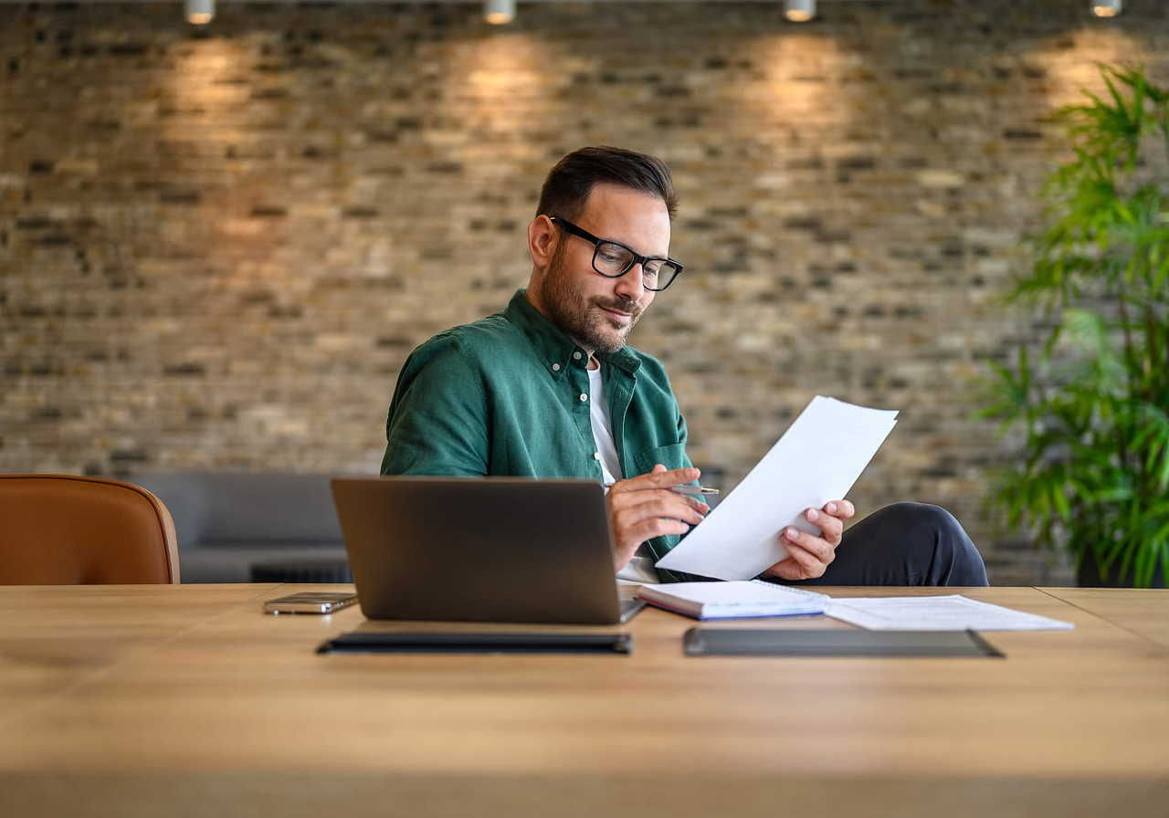 A man in a green shirt and glasses reviews documents at a wooden desk with a laptop, set against a modern brick wall and greenery.